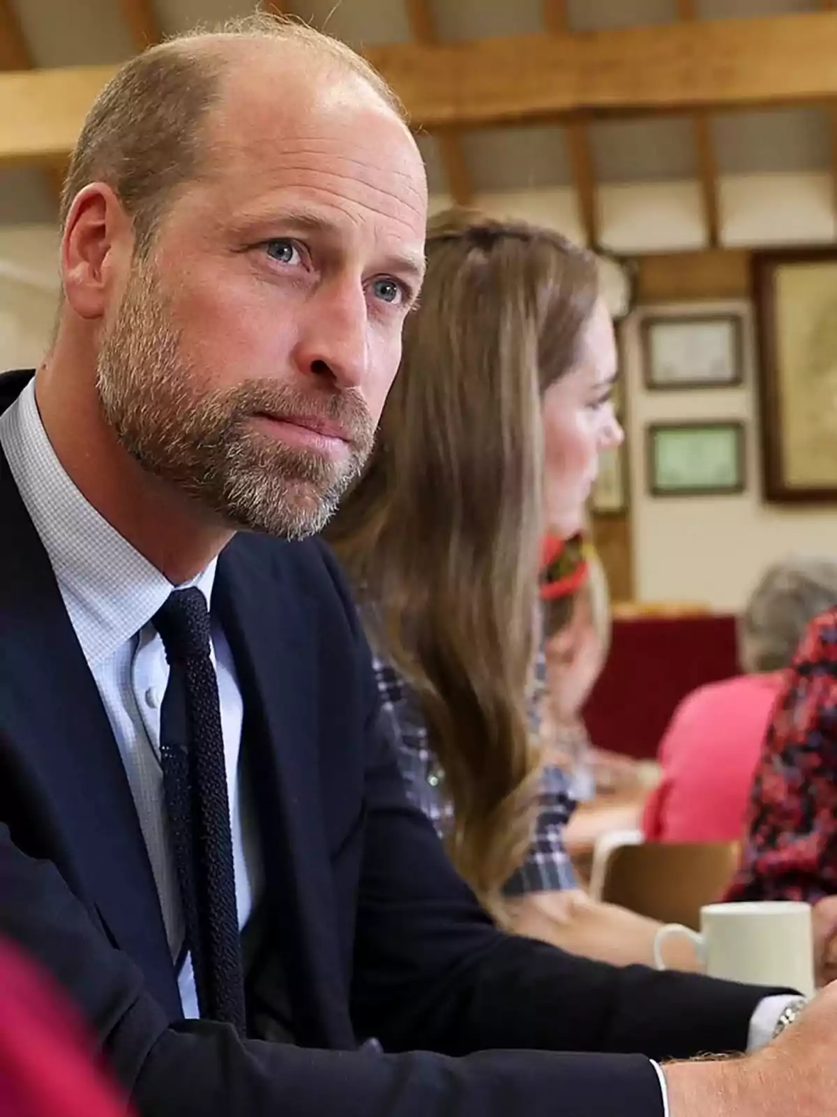 El príncipe Guillermo de traje oscuro sentado en una mesa en un ambiente interior con otras personas al fondo