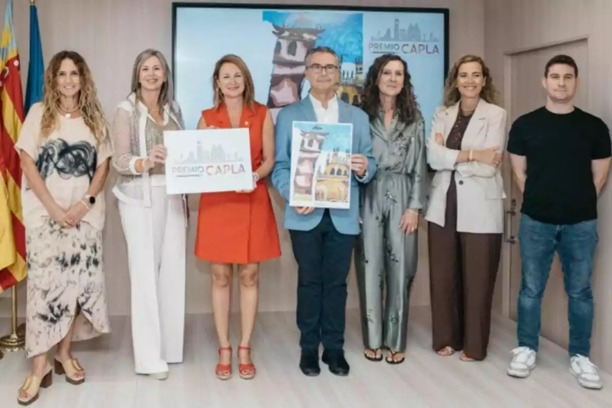Siete personas posan en una sala junto a una pantalla y banderas, dos de ellas sostienen carteles relacionados con un premio llamado CAPLA Siete personas posan en una sala junto a una pantalla y banderas, dos de ellas sostienen carteles relacionados con un premio llamado CAPLA
