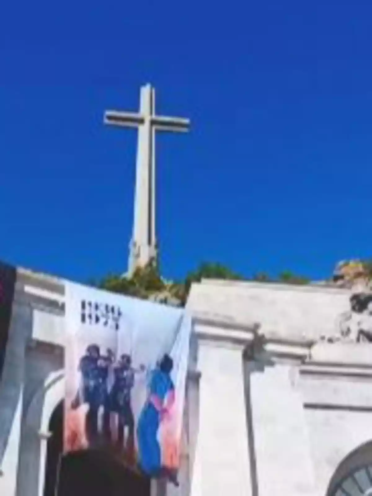 Cruz monumental sobre un edificio de piedra con una pancarta colgada en la fachada y cielo azul de fondo