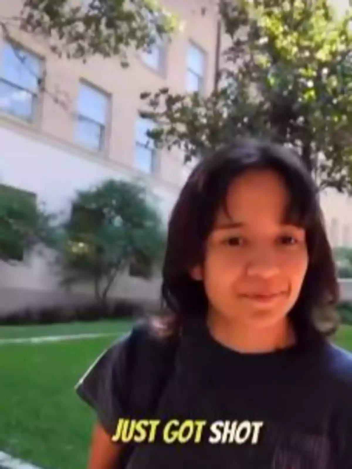 Persona joven de cabello oscuro mirando a la cámara en un jardín con un edificio al fondo y el texto just got shot en la parte inferior