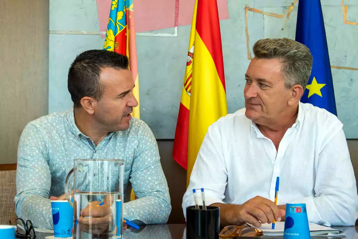 Dos hombres conversan sentados en una mesa con banderas oficiales de fondo y materiales de oficina sobre la mesa Dos hombres conversan sentados en una mesa con banderas oficiales de fondo y materiales de oficina sobre la mesa