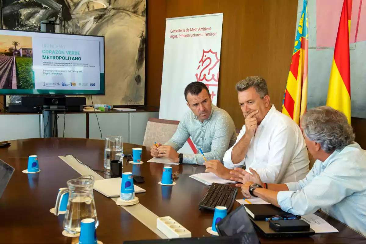 Tres hombres conversan en una sala de reuniones con una mesa redonda, papeles, ordenadores y vasos de agua, mientras al fondo se ve una pantalla con una presentación sobre un proyecto verde y varias banderas oficiales
