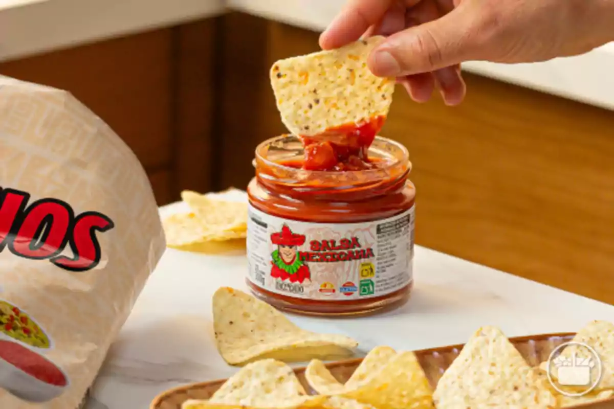 Una mano sostiene un totopo sumergido en un frasco de salsa mexicana junto a una bolsa de nachos y varios totopos sobre la mesa Una mano sostiene un totopo sumergido en un frasco de salsa mexicana junto a una bolsa de nachos y varios totopos sobre la mesa