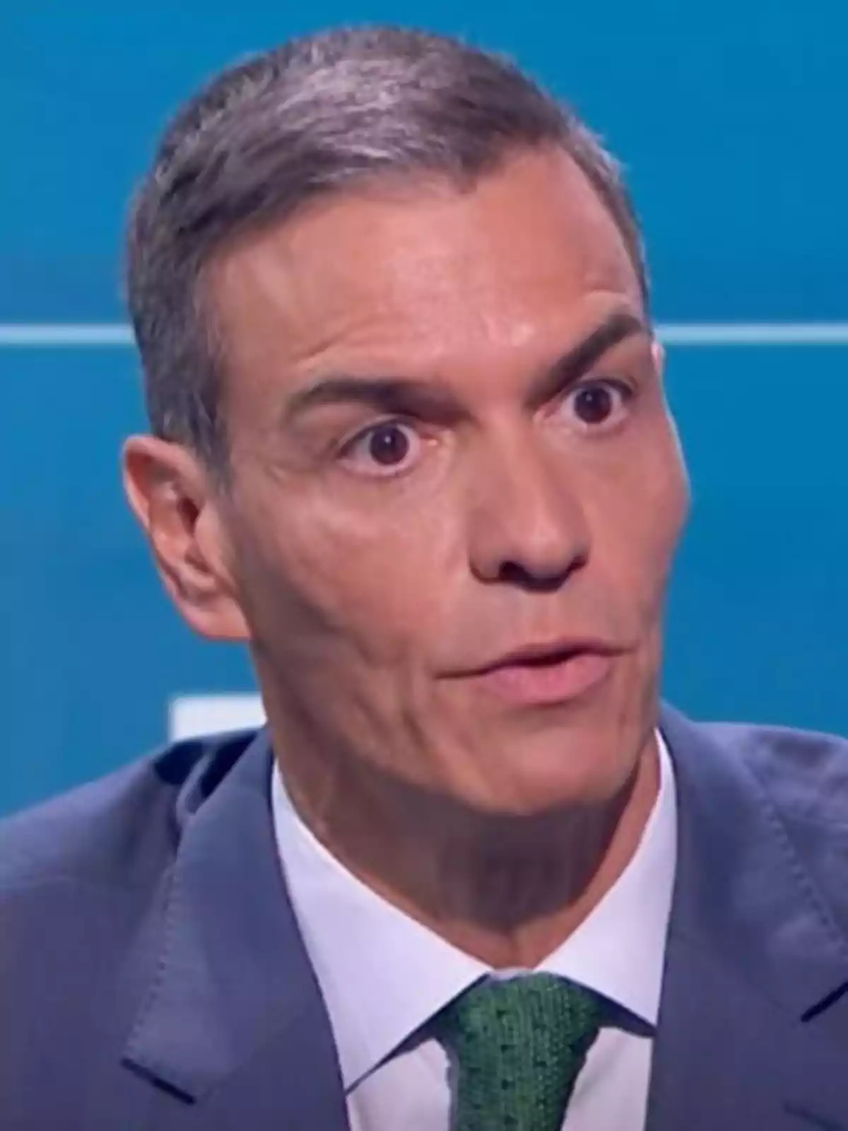 Hombre de cabello corto y canoso vistiendo traje azul, camisa blanca y corbata verde, con expresión seria frente a un fondo azul Hombre de cabello corto y canoso vistiendo traje azul, camisa blanca y corbata verde, con expresión seria frente a un fondo azul