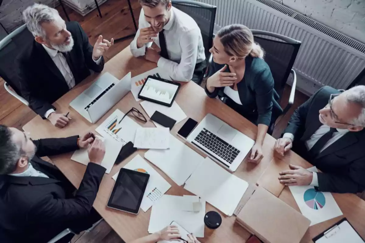 Grupo de personas en una reunión de trabajo alrededor de una mesa con computadoras portátiles, tabletas, documentos y gráficos