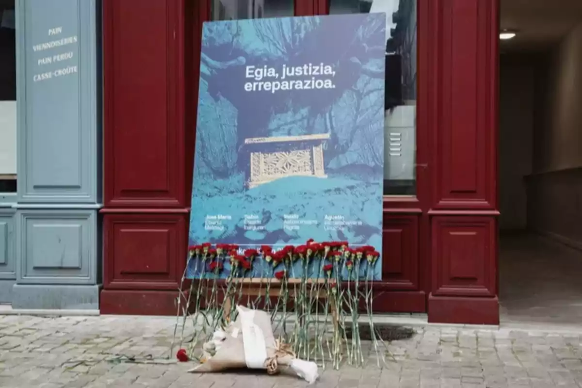 Cartel azul con letras blancas y flores rojas apoyadas en la base, colocado frente a una puerta roja en una calle de adoquines