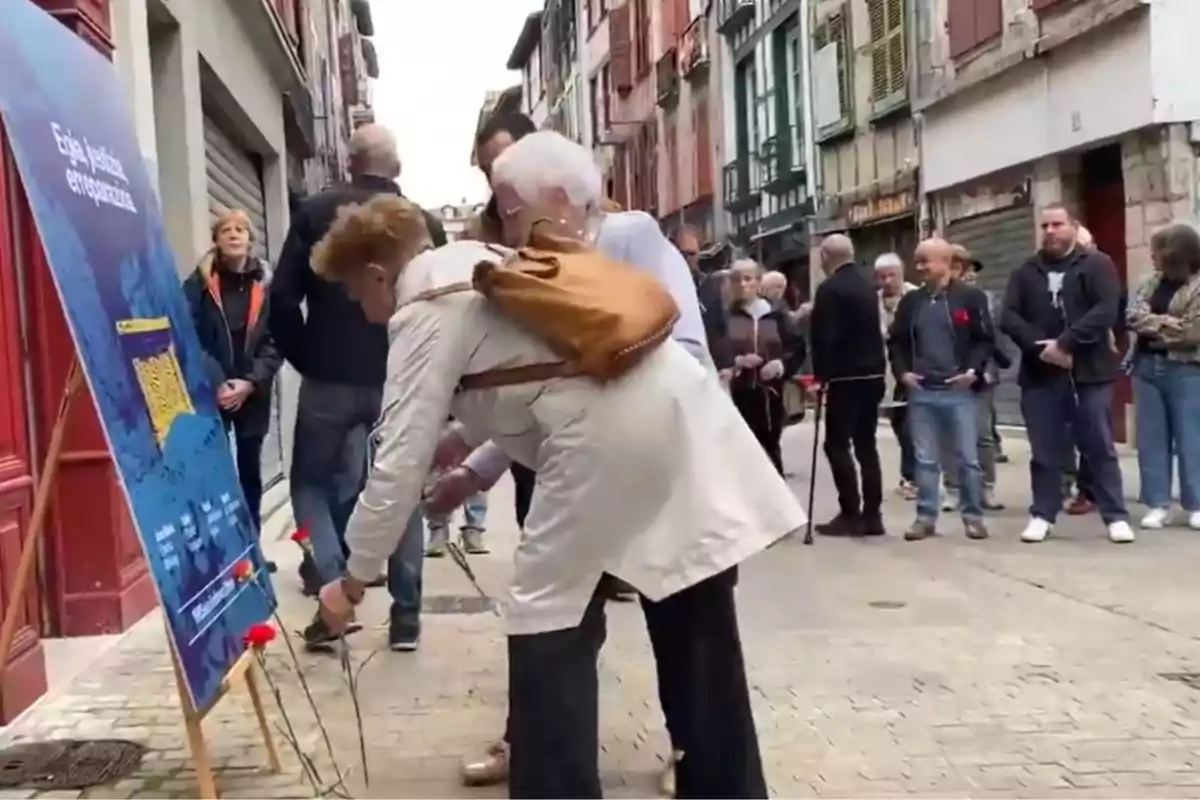 Personas en una calle colocando flores rojas en un cartel mientras otras esperan en fila observando la escena