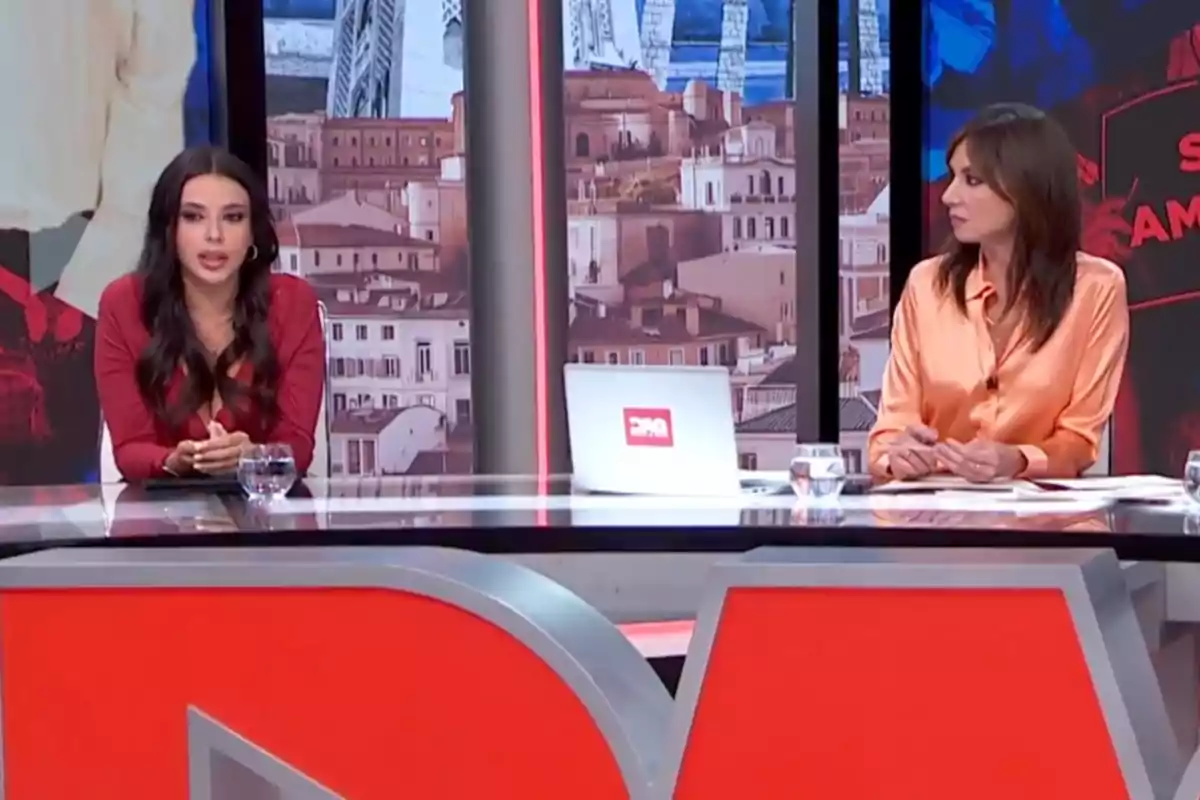 Dos mujeres sentadas en un set de televisión con fondo de ciudad y una mesa con vasos de agua y una laptop