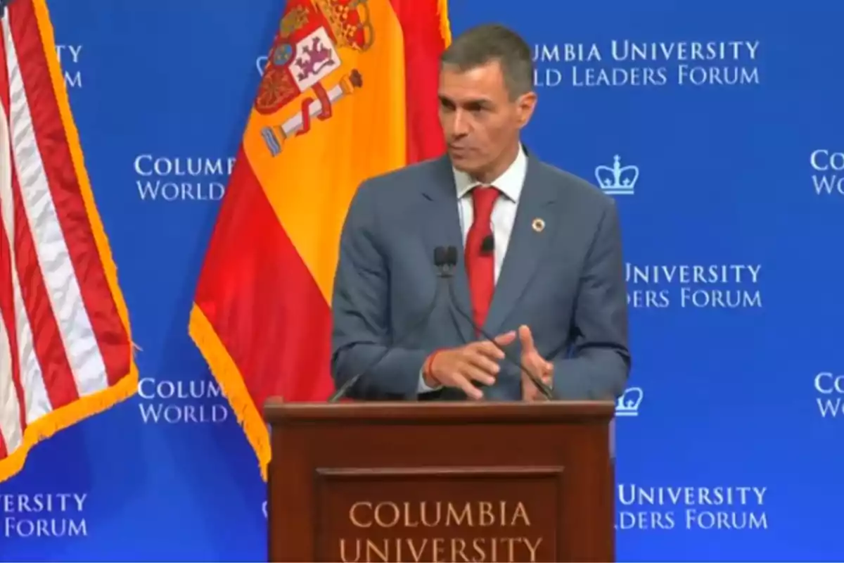 Hombre con traje azul y corbata roja hablando en un podio de Columbia University con las banderas de España y Estados Unidos de fondo