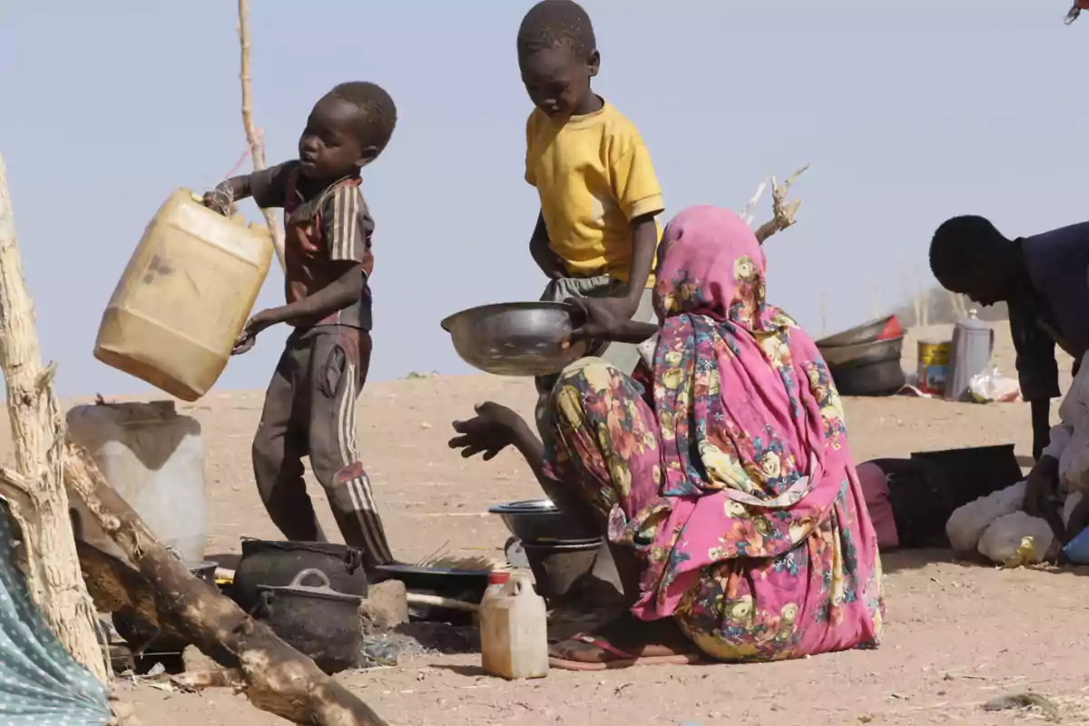 Una mujer y tres niños en un entorno árido realizan tareas domésticas al aire libre usando recipientes de plástico y metal