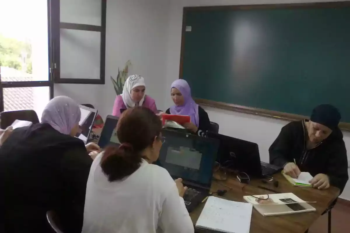 Cinco mujeres trabajan en una mesa con computadoras portátiles y cuadernos en un aula frente a un pizarrón verde