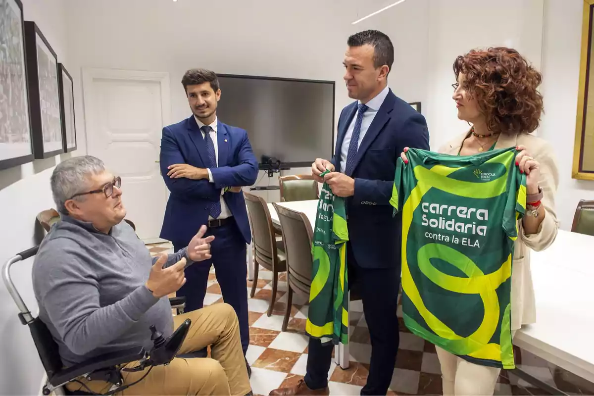 Cuatro personas conversan en una oficina mientras dos de ellas sostienen camisetas verdes con el texto carrera solidaria contra la ELA