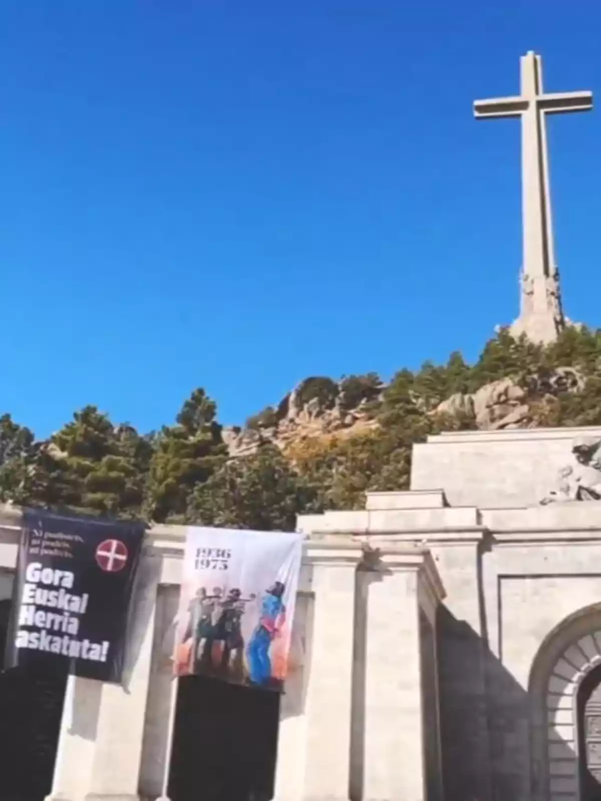Cruz monumental sobre una colina con dos pancartas colgadas en la entrada de un edificio de piedra