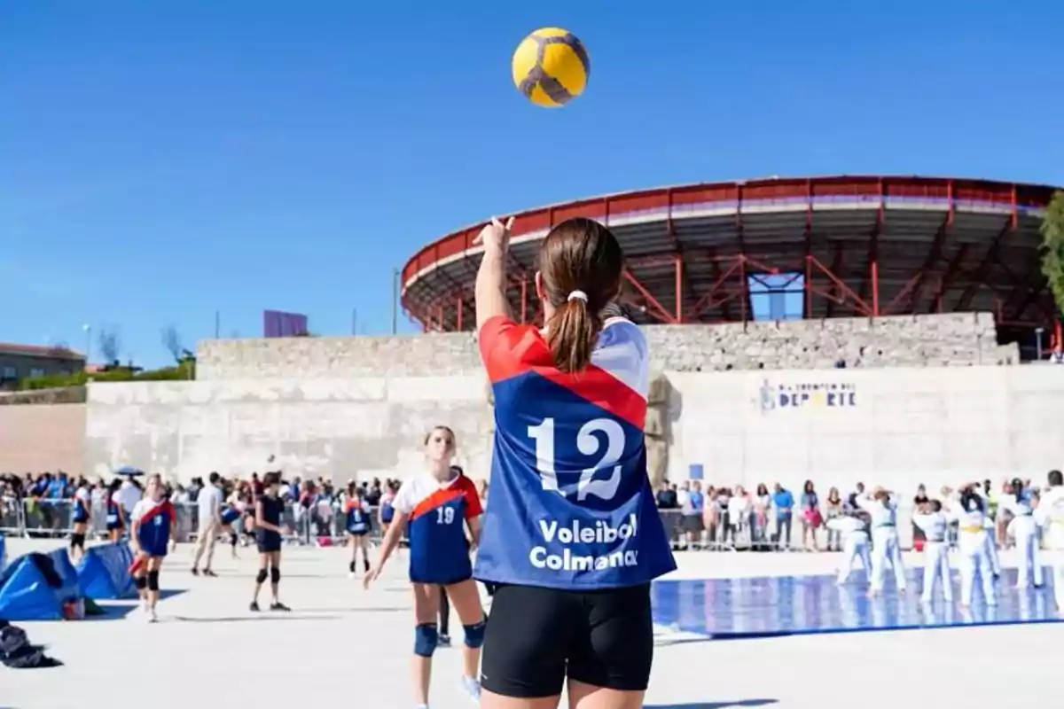 Jugadora de voleibol con el uniforme azul, rojo y blanco lanzando el balón en una cancha al aire libre con público y otros deportistas al fondo