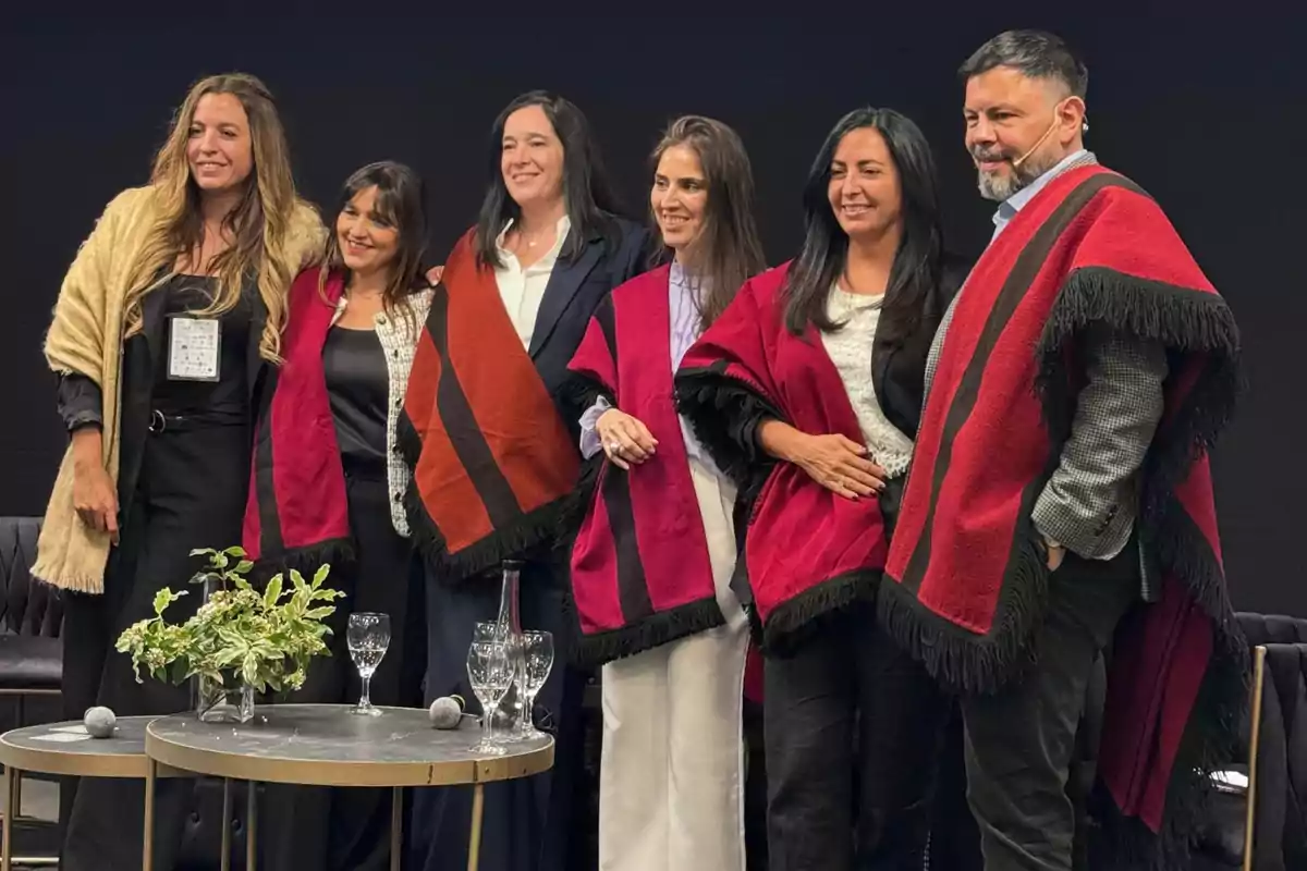 Seis personas posan juntas en un evento, cinco de ellas llevan ponchos tradicionales de colores rojo y marrón, mientras que una mujer lleva un chal beige, al frente hay una mesa con copas, una botella de agua y un arreglo floral. Seis personas posan juntas en un evento, cinco de ellas llevan ponchos tradicionales de colores rojo y marrón, mientras que una mujer lleva un chal beige, al frente hay una mesa con copas, una botella de agua y un arreglo floral.