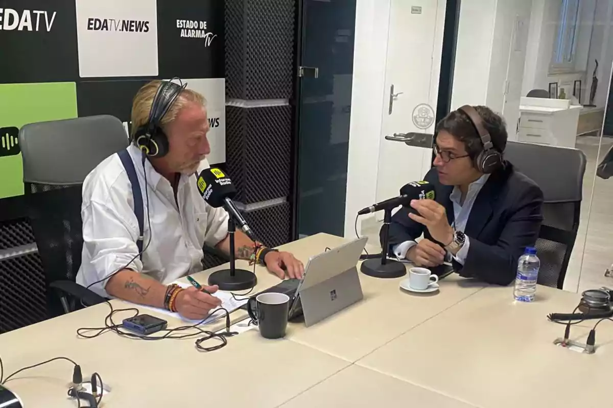 Dos hombres con auriculares y micrófonos conversan en una mesa de estudio de radio, uno de ellos toma notas mientras el otro gesticula, ambos rodeados de tazas, una botella de agua y una tablet, con un fondo de paneles acústicos y logotipos de un canal de noticias. Dos hombres con auriculares y micrófonos conversan en una mesa de estudio de radio, uno de ellos toma notas mientras el otro gesticula, ambos rodeados de tazas, una botella de agua y una tablet, con un fondo de paneles acústicos y logotipos de un canal de noticias.