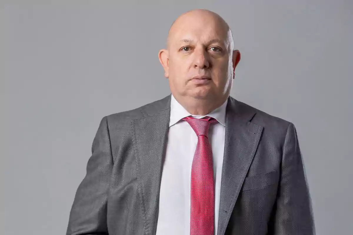 Hombre calvo con traje gris, camisa blanca y corbata rosa posando frente a un fondo gris