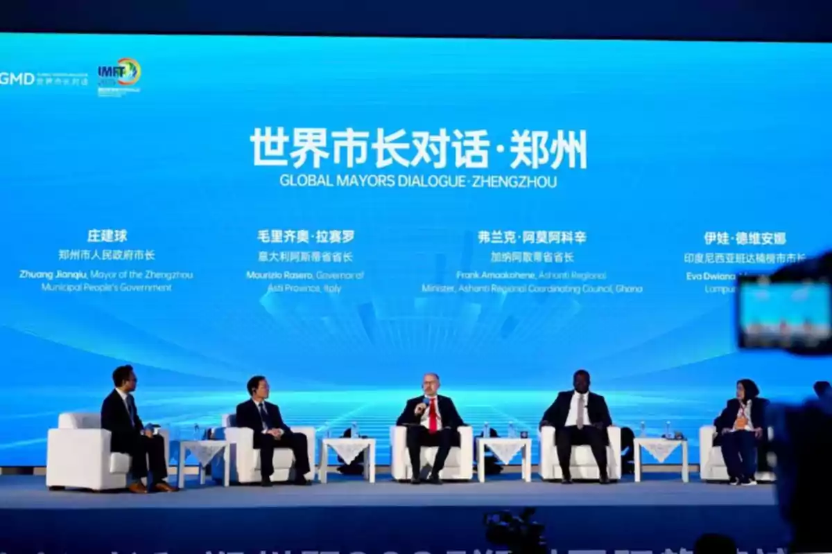 Cinco personas sentadas en sillones blancos participan en un panel de discusión en un escenario con fondo azul que muestra el título Global Mayors Dialogue Zhengzhou y nombres de los participantes en inglés y chino