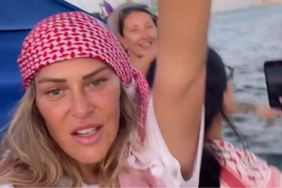 Mujer con pañuelo rojo y blanco en la cabeza levantando el brazo mientras sonríe en un ambiente al aire libre cerca del agua