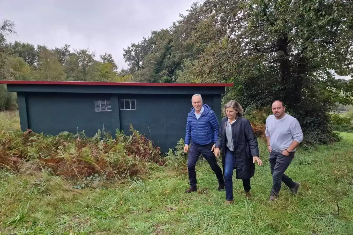 Tres personas caminan por un terreno cubierto de césped y helechos frente a un edificio pequeño de color oscuro con techo rojo y árboles al fondo Tres personas caminan por un terreno cubierto de césped y helechos frente a un edificio pequeño de color oscuro con techo rojo y árboles al fondo