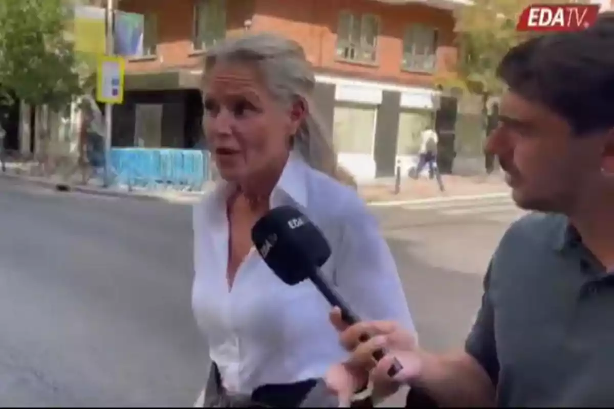 Una mujer de cabello canoso y camisa blanca es entrevistada en la calle por un reportero que sostiene un micrófono de EDA TV Una mujer de cabello canoso y camisa blanca es entrevistada en la calle por un reportero que sostiene un micrófono de EDA TV
