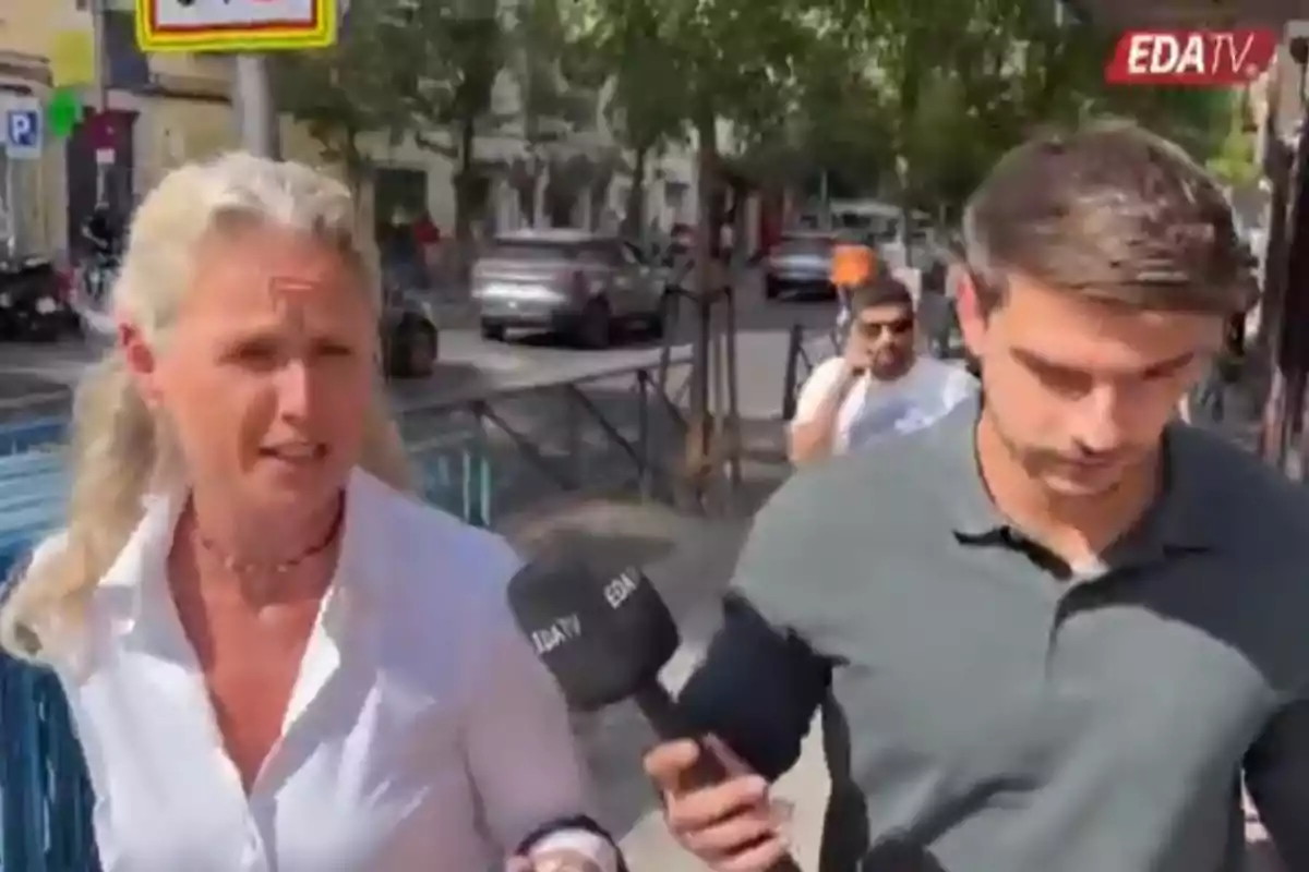 Una mujer de cabello rubio y un hombre con camisa gris caminan por una acera mientras un reportero sostiene un micrófono con el logo de EDATV Una mujer de cabello rubio y un hombre con camisa gris caminan por una acera mientras un reportero sostiene un micrófono con el logo de EDATV