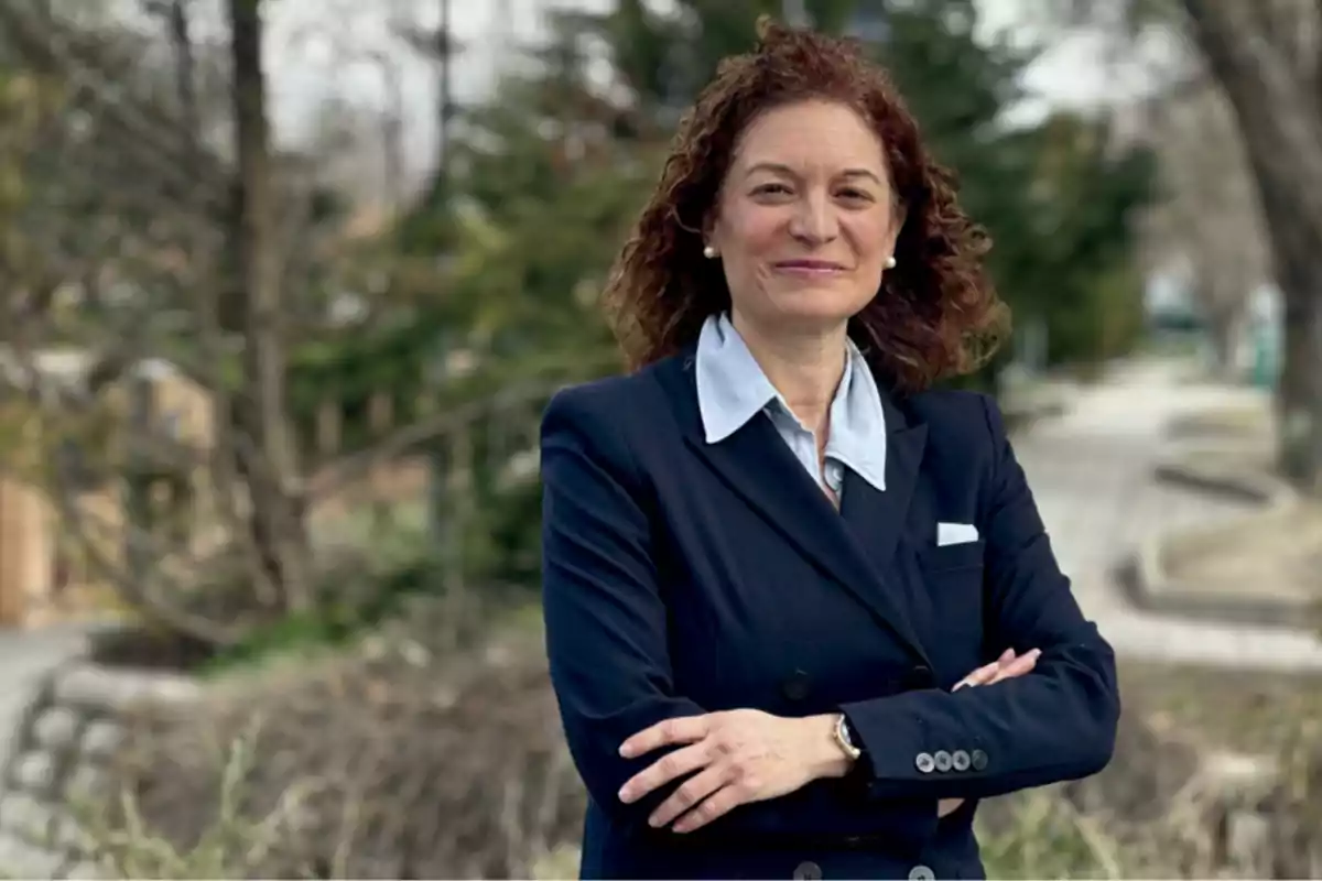 Mujer de cabello rizado y castaño con saco azul marino y camisa clara posando con los brazos cruzados en un entorno al aire libre con árboles y sendero al fondo Mujer de cabello rizado y castaño con saco azul marino y camisa clara posando con los brazos cruzados en un entorno al aire libre con árboles y sendero al fondo