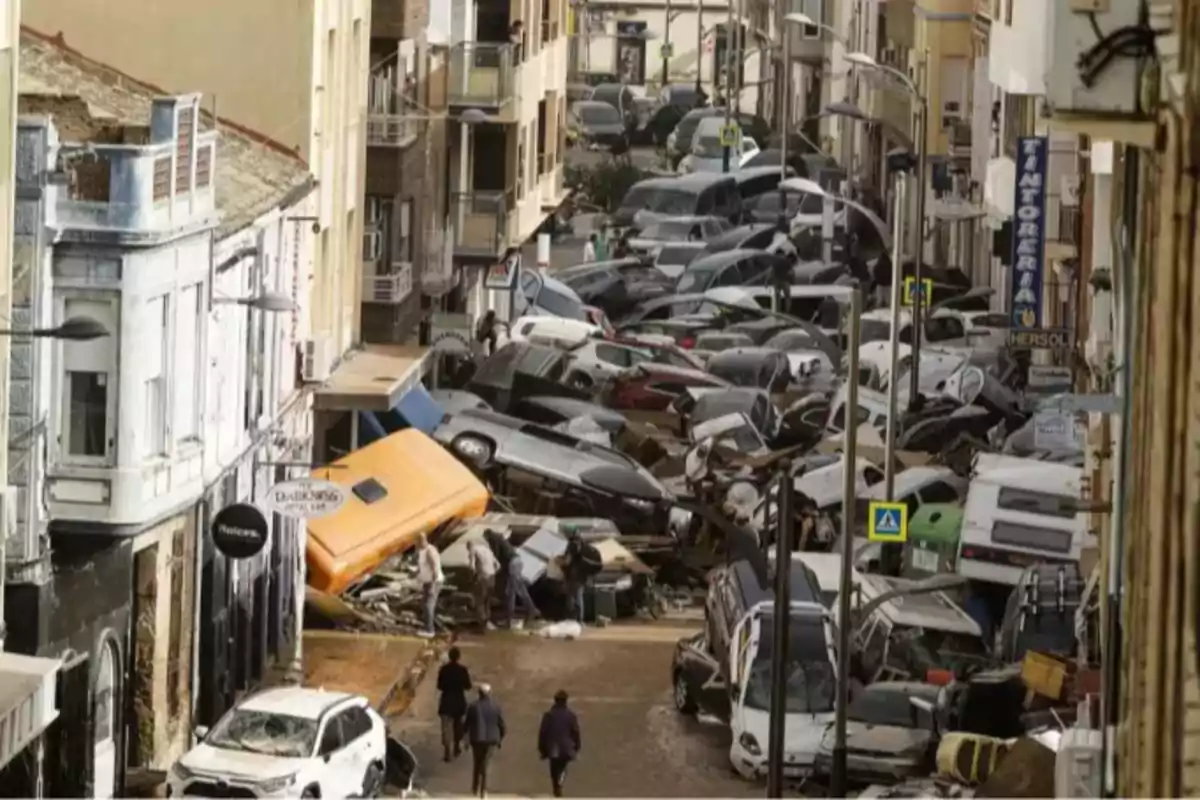 Calle llena de autos apilados y dañados tras una inundación con personas caminando entre los escombros Calle llena de autos apilados y dañados tras una inundación con personas caminando entre los escombros
