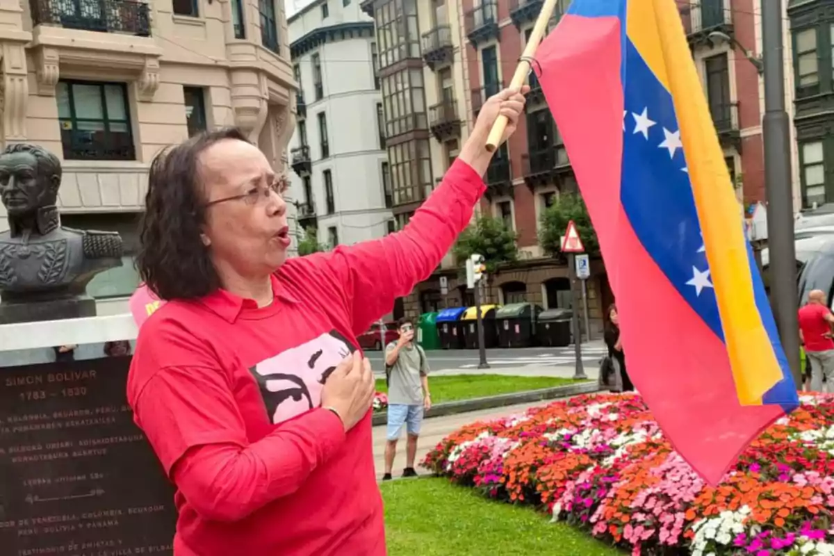 Mujer con camiseta roja sostiene una bandera de Venezuela en una plaza con flores y edificios al fondo Mujer con camiseta roja sostiene una bandera de Venezuela en una plaza con flores y edificios al fondo
