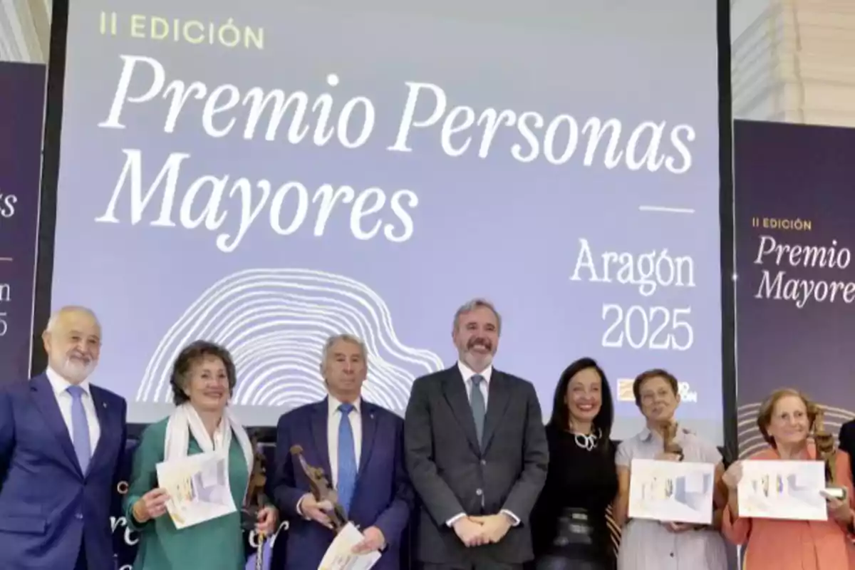 Grupo de personas sonrientes posando con diplomas y trofeos frente a un cartel que anuncia la II Edición del Premio Personas Mayores en Aragón 2025 Grupo de personas sonrientes posando con diplomas y trofeos frente a un cartel que anuncia la II Edición del Premio Personas Mayores en Aragón 2025