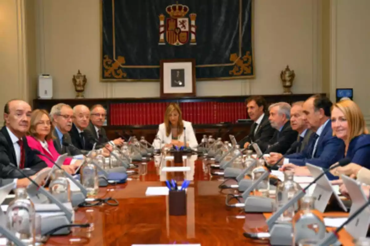 Un grupo de personas en una sala de reuniones sentadas alrededor de una mesa larga con micrófonos y documentos, al fondo una bandera con el escudo de España y estanterías con libros.