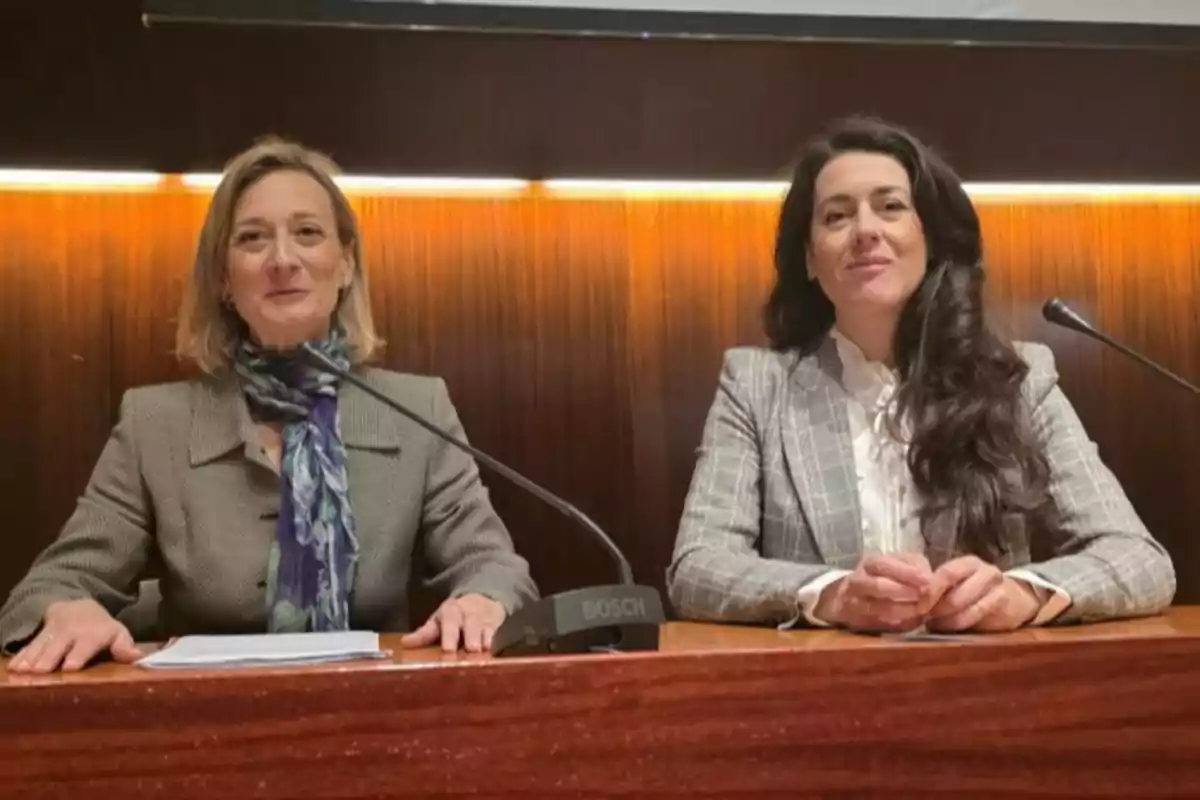 Dos mujeres sentadas en una mesa de madera durante una conferencia, ambas con chaquetas formales y micrófonos frente a ellas.