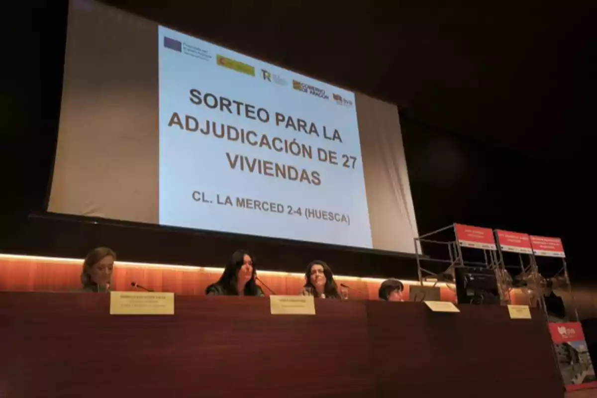 Cuatro personas sentadas en una mesa frente a una pantalla grande que anuncia un sorteo para la adjudicación de 27 viviendas en Huesca