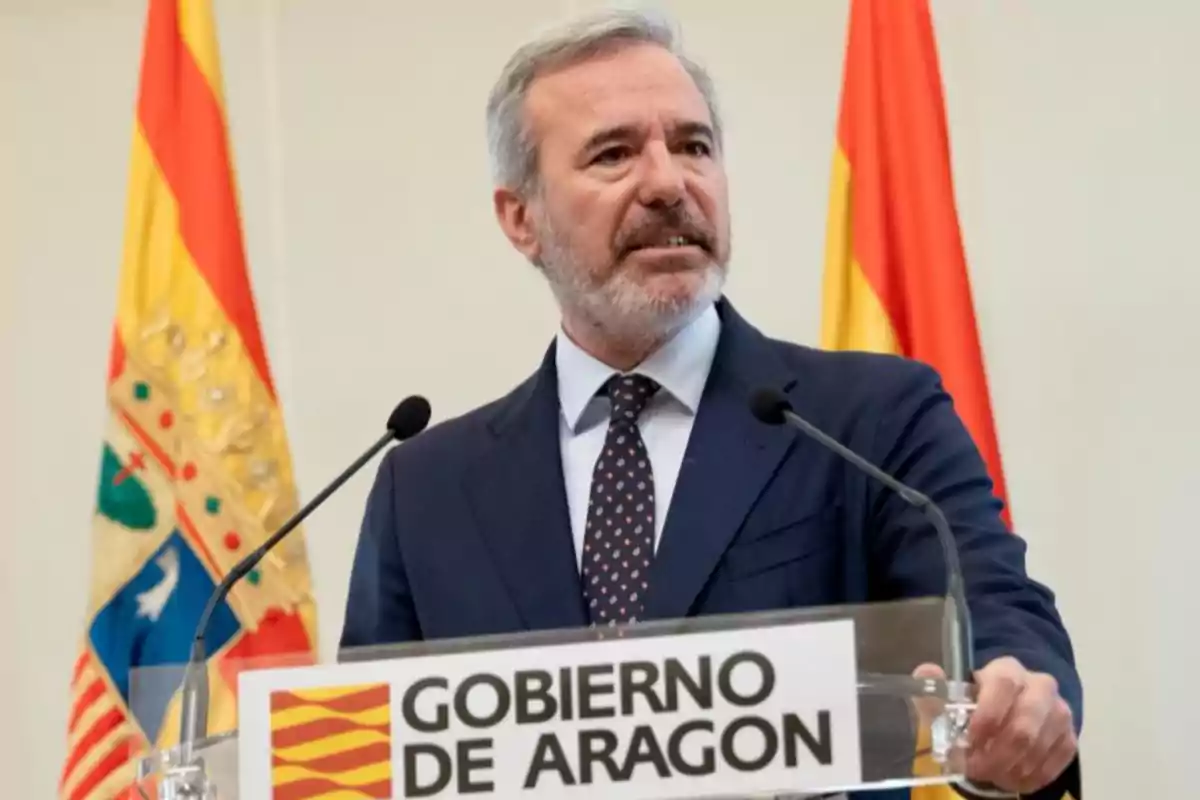 Hombre de cabello canoso y barba hablando en un atril con el letrero Gobierno de Aragón y banderas de fondo