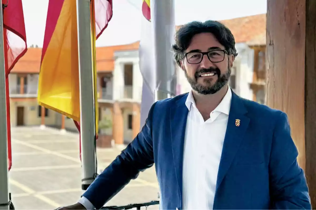 Hombre con gafas y barba sonríe mientras posa con un saco azul en un balcón con banderas y un edificio antiguo de fondo Hombre con gafas y barba sonríe mientras posa con un saco azul en un balcón con banderas y un edificio antiguo de fondo