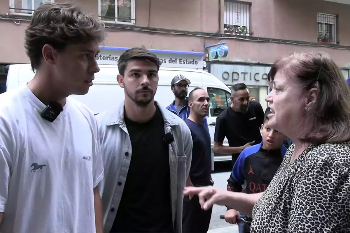 Un grupo de personas conversa en la calle frente a una óptica, una mujer señala mientras dos jóvenes la escuchan atentamente y varias personas observan la escena al fondo.
