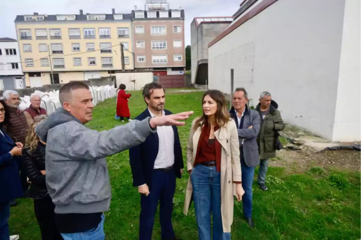 Un grupo de personas conversa y observa un terreno al aire libre junto a un edificio, mientras una persona señala hacia el frente