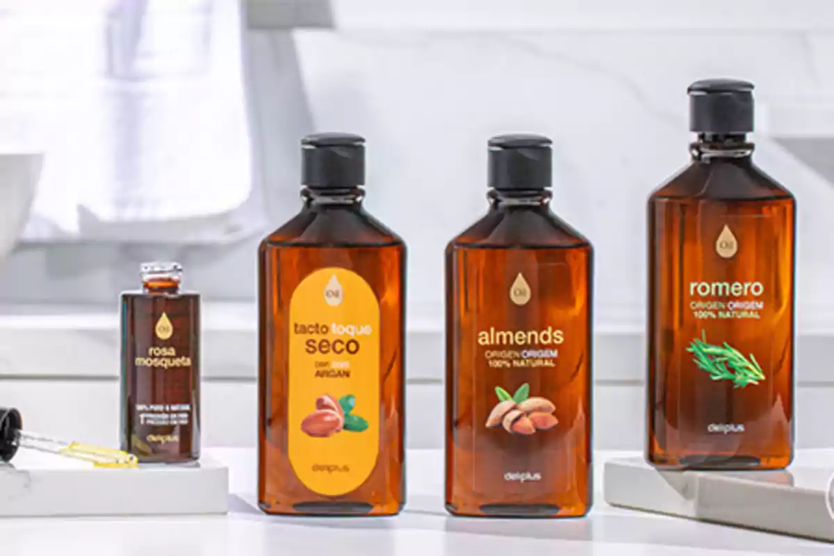 Cuatro frascos de aceites naturales de la marca Deliplus están alineados sobre una superficie blanca en un baño, cada uno con diferentes etiquetas y colores representando rosa mosqueta, tacto seco con argán, almendras y romero