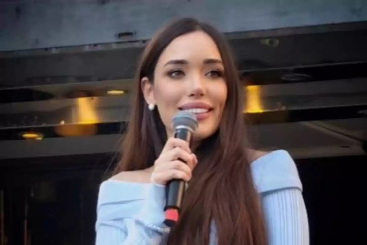Mujer de cabello largo y lacio usando un suéter azul claro y sosteniendo un micrófono mientras sonríe