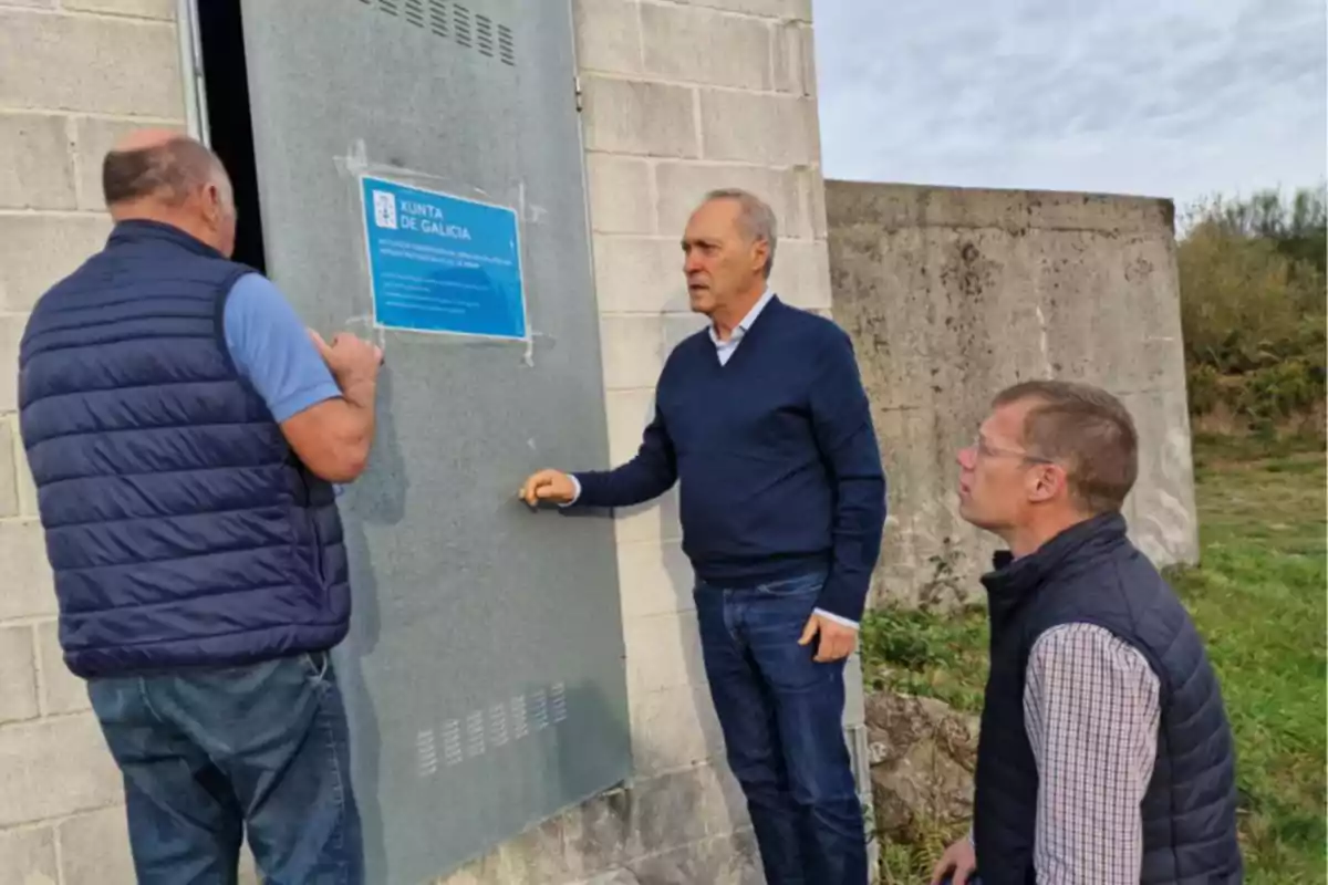 Tres hombres con chalecos y suéteres conversan frente a una puerta metálica en un edificio de bloques, al aire libre, junto a un cartel azul de la Xunta de Galicia.
