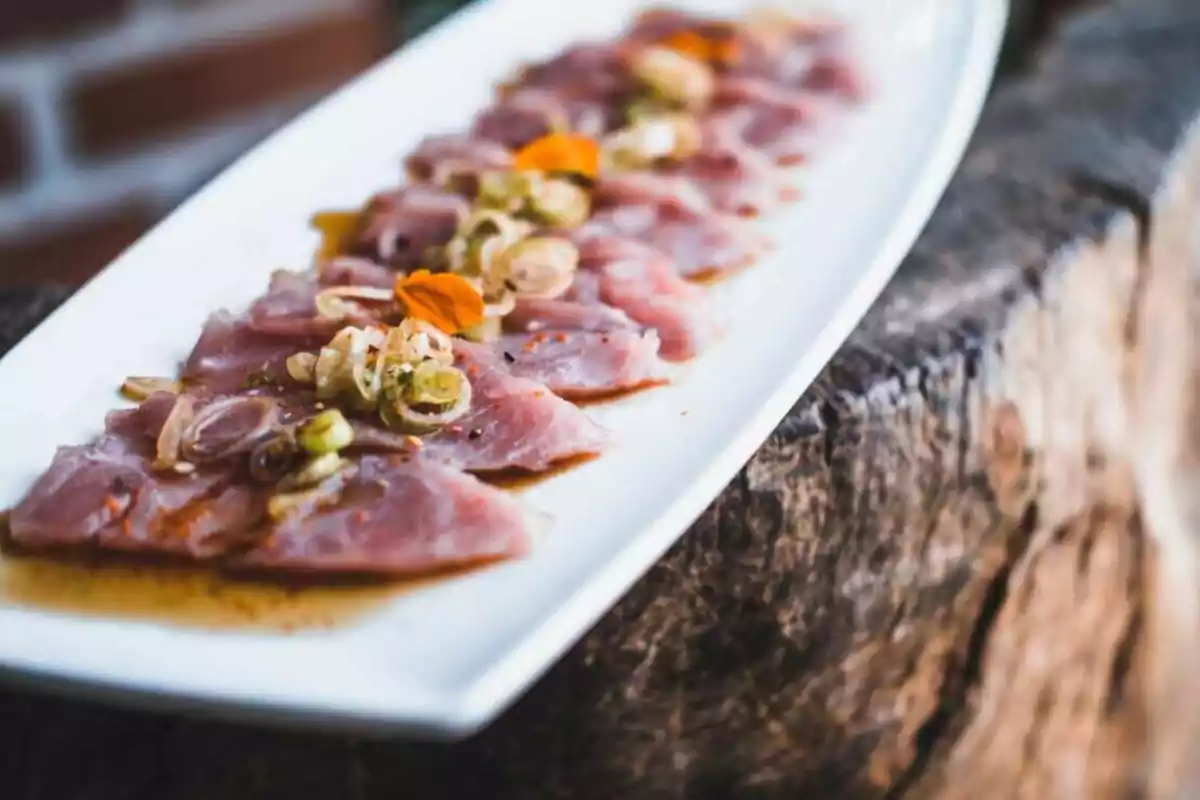 Plato rectangular con láminas de pescado crudo decoradas con cebolla y flores comestibles sobre una base de salsa Plato rectangular con láminas de pescado crudo decoradas con cebolla y flores comestibles sobre una base de salsa