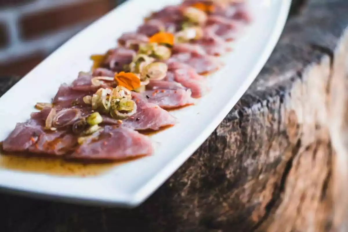 Plato rectangular con láminas de pescado crudo decoradas con cebolla, hierbas y pétalos naranjas sobre una base de madera rústica Plato rectangular con láminas de pescado crudo decoradas con cebolla, hierbas y pétalos naranjas sobre una base de madera rústica