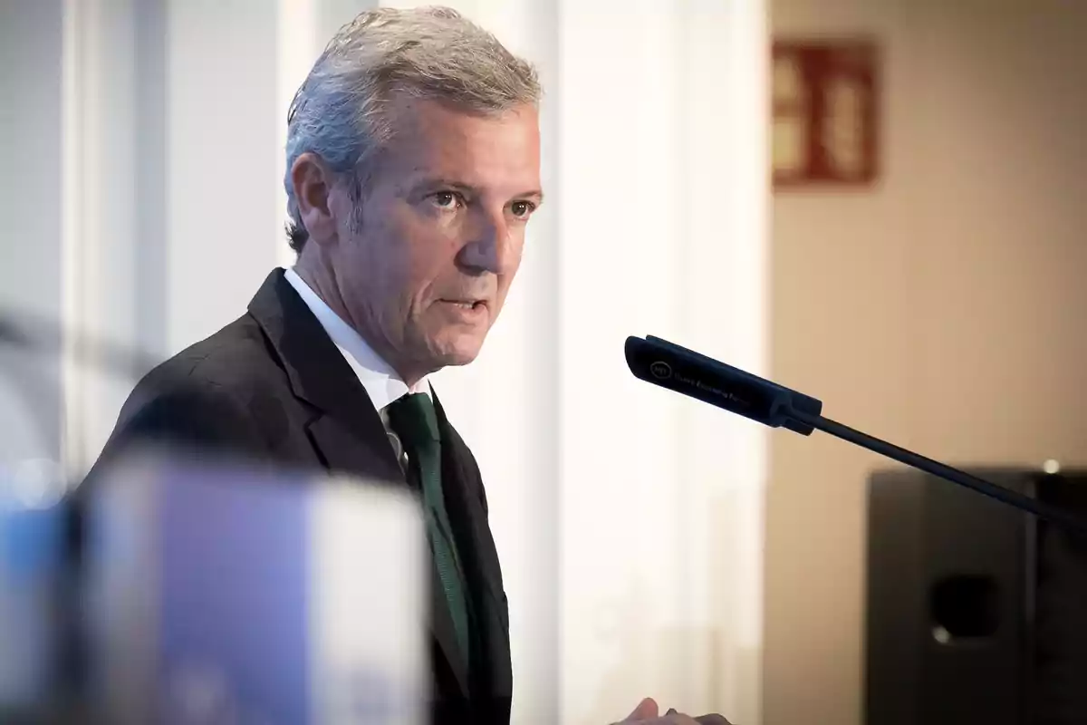 Hombre de cabello canoso con traje oscuro hablando frente a un micrófono en un entorno profesional