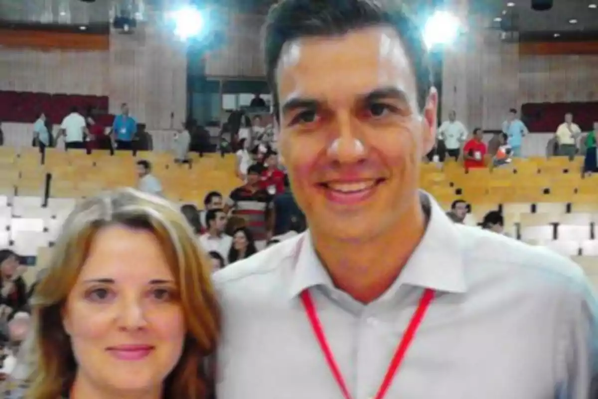Dos personas posan para una foto en un auditorio con asientos de madera y varias personas al fondo. Dos personas posan para una foto en un auditorio con asientos de madera y varias personas al fondo.