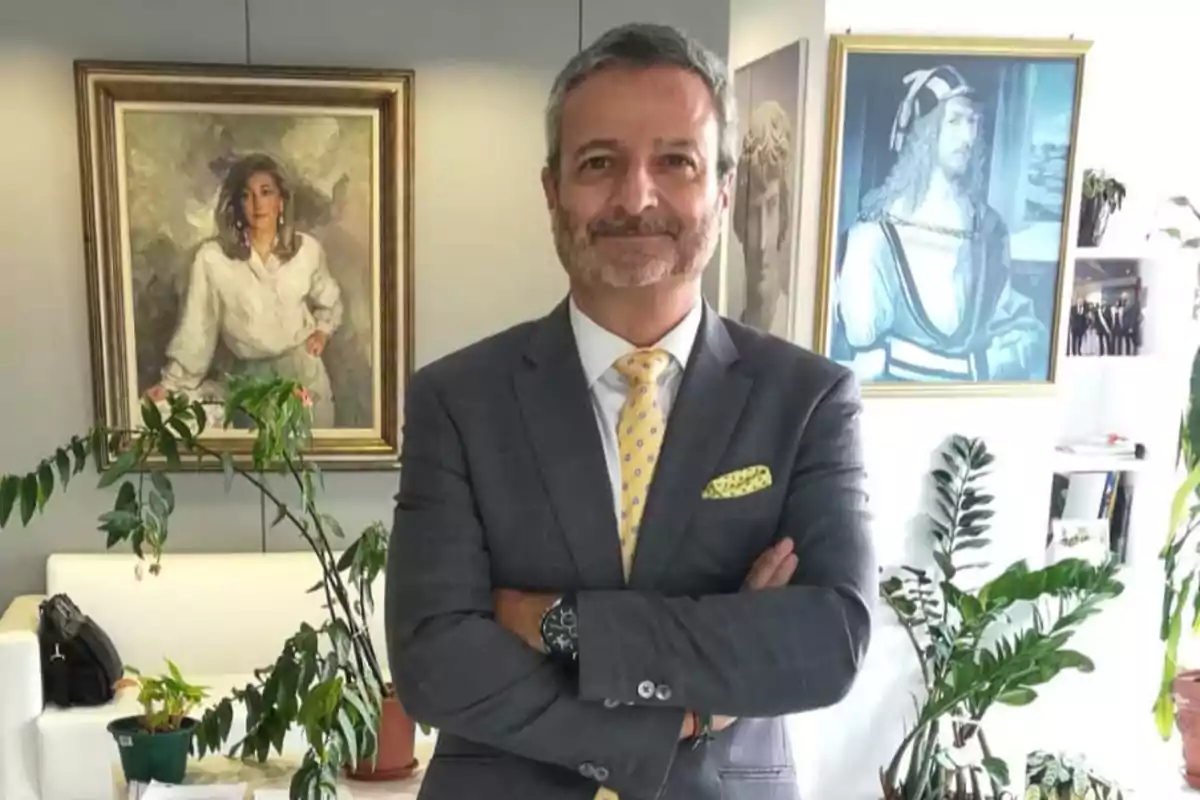 Hombre de traje gris con corbata amarilla posando con los brazos cruzados en una oficina decorada con plantas y cuadros al fondo