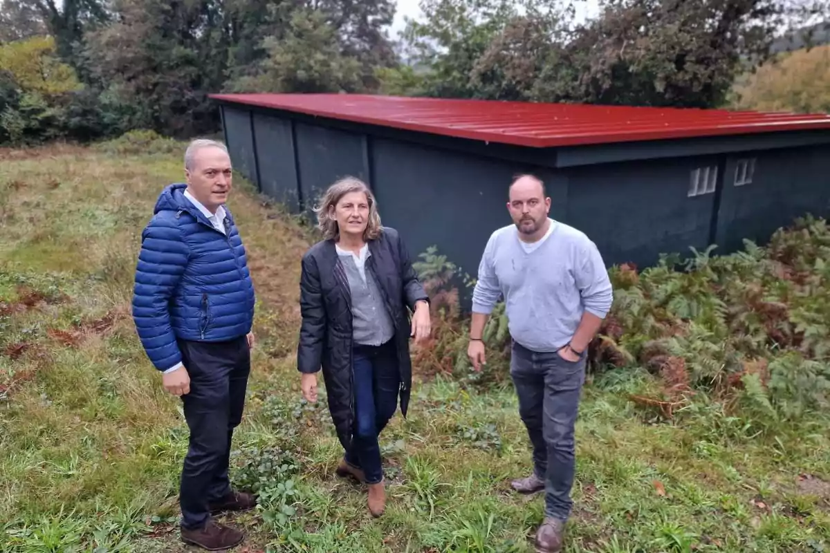 Tres personas posan de pie sobre césped frente a un edificio de techo rojo rodeado de vegetación y árboles.