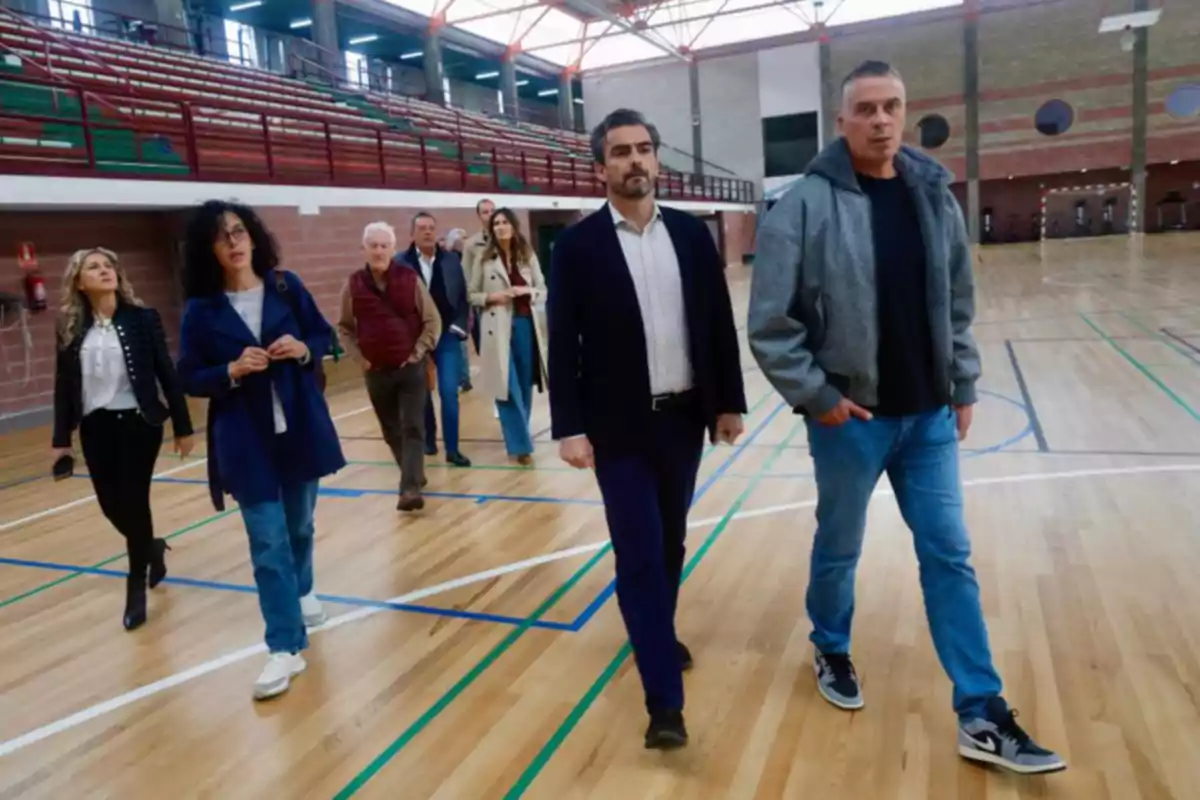 Un grupo de personas camina por el interior de un gimnasio con piso de madera y gradas al fondo Un grupo de personas camina por el interior de un gimnasio con piso de madera y gradas al fondo