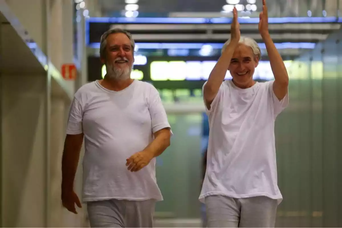 Dos personas adultas sonrientes caminan por un pasillo iluminado, una de ellas aplaude mientras la otra avanza con expresión alegre, ambas visten ropa cómoda de color blanco y gris. Dos personas adultas sonrientes caminan por un pasillo iluminado, una de ellas aplaude mientras la otra avanza con expresión alegre, ambas visten ropa cómoda de color blanco y gris.