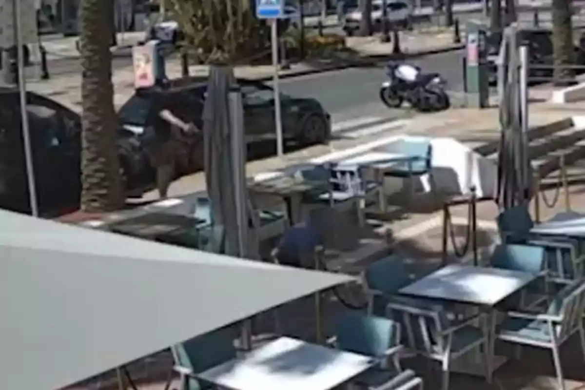 Terraza de cafetería con mesas y sillas vacías, una persona en el suelo y otra junto a un coche estacionado en la acera, al fondo se ve una motocicleta sobre la banqueta. Terraza de cafetería con mesas y sillas vacías, una persona en el suelo y otra junto a un coche estacionado en la acera, al fondo se ve una motocicleta sobre la banqueta.