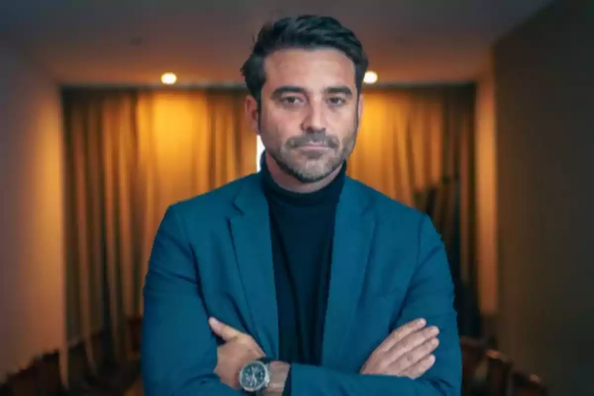 Hombre de cabello corto y barba con expresión seria, vestido con saco azul y suéter negro, de brazos cruzados en una habitación con cortinas y luz cálida de fondo